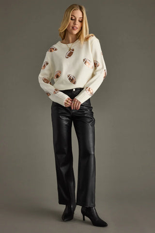 Woman wearing a cream sweater with patterned design and black pants on a gray background