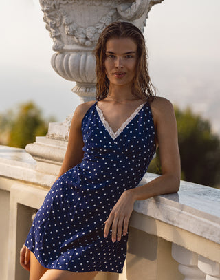 Woman in a blue polka dot dress standing outdoors near classical architecture.