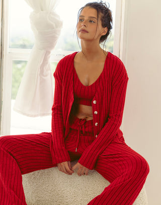 Woman lounging on a cream textured sofa wearing a cozy red ribbed knit lounge set, including a cropped tank top, drawstring pants, and an open cardigan, with natural light streaming through white curtains behind her.