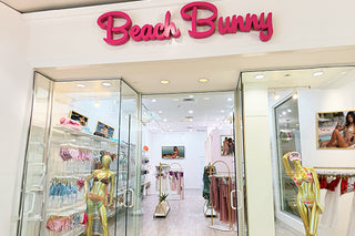 Beach Bunny boutique storefront with a large pink script logo above glass doors, interior racks of swimwear and clothing, and gold mannequins displaying swimwear.