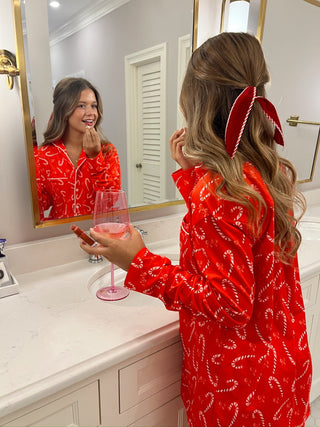 Woman in a red robe with candy cane pattern applying makeup in front of a mirror.