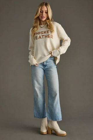Woman wearing a sweater with 'Whiskey Weather' text and jeans on a gray background