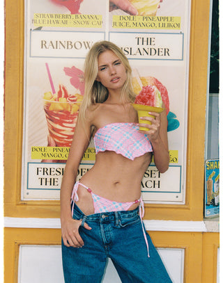 A woman stands in front of a colorful ice cream and drink sign, wearing a pink and white plaid bikini top and matching bottoms with loose blue jeans.